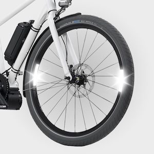 May include: Close-up of a white electric bicycle front wheel. The black tire has a reflective strip and the wheel has a disc brake. A black battery pack is mounted on the frame. The bike has a headlight and a black fender.