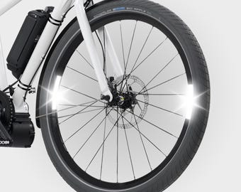 May include: Close-up of a white electric bicycle front wheel. The black tire has a reflective strip and the wheel has a disc brake. A black battery pack is mounted on the frame. The bike has a headlight and a black fender.