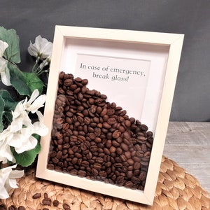 May include: A white wooden frame with a glass front filled with coffee beans. The frame has a white background with the text "In case of emergency, break glass!" printed on it.