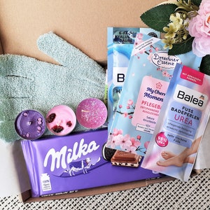 Puede incluir: Una caja de regalo llena de productos de ba&ntilde;o y cuerpo, que incluyen una barra de chocolate morada, un guante exfoliante verde, tres velas peque&ntilde;as y un paquete de producto de ba&ntilde;o rosa y azul. La caja est&aacute; forrada con papel de seda blanco y decorada con flores rosas.