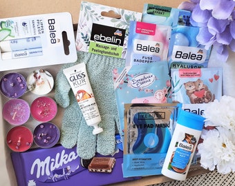 Könnte beinhalten: Eine Geschenkbox gefüllt mit verschiedenen Beauty- und Pflegeprodukten, darunter Gesichtsmasken, Handcreme, Lippenbalsam, Badebomben und Kerzen. Die Box enthält auch eine Tafel Schokolade und ein Paar grüne Peelinghandschuhe.