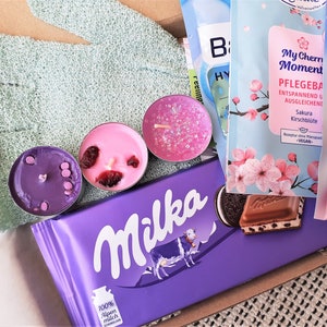 Puede incluir: Una barra de chocolate Milka morada con una vaca en la parte delantera, una vela morada, rosa y blanca, y un paquete de crema para pies rosa y azul con el texto "Bale Fu&szlig; Badepeeling Urea 95% Urea" y "My Cherry Moment Pflegebad Entspannend und Ausgleichend Sakura Kirschbl&uuml;te"