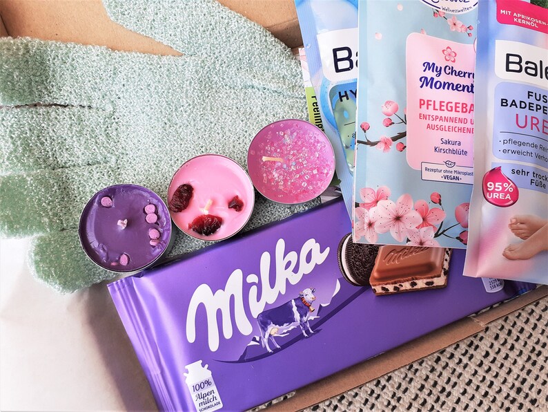 Puede incluir: Una barra de chocolate Milka morada con una vaca en la parte delantera, una vela morada, rosa y blanca, y un paquete de crema para pies rosa y azul con el texto "Bale Fu&szlig; Badepeeling Urea 95% Urea" y "My Cherry Moment Pflegebad Entspannend und Ausgleichend Sakura Kirschbl&uuml;te"
