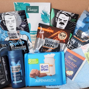 Puede incluir: Una caja llena de varios productos de aseo para hombres, incluyendo mascarillas faciales, gel de ducha, bálsamo labial y una barra de chocolate. Los productos son de las marcas Balea, Kneipp y Ritter Sport. La caja también incluye una toallita azul y una pequeña bolsa de té.
