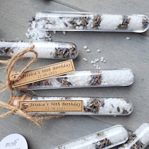 May include: Small glass tubes filled with white bath salts and dried lavender flowers. Each tube has a brown paper tag with the text "Jessica's 30th Birthday 22.08.2023".