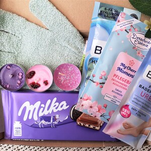 Puede incluir: Una caja de regalo llena de diversos productos de ba&ntilde;o y cuerpo, incluyendo una barra de chocolate morada, un par de guantes de ba&ntilde;o de color turquesa, tres velas arom&aacute;ticas rosas y moradas, y tres paquetes de productos de ba&ntilde;o. Los productos de ba&ntilde;o est&aacute;n etiquetados como "6-fach Hyaluron S&auml;ure", "Dresdner Essenz", "My Cherry Moment Pflegebad" y "Balea Fuss Badeperlen Urea".