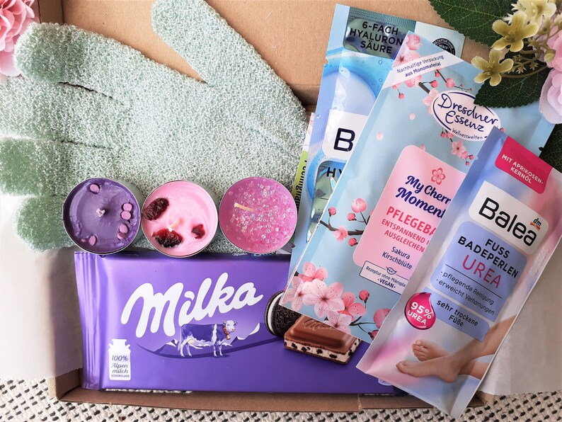Puede incluir: Una caja de regalo llena de diversos productos de ba&ntilde;o y cuerpo, incluyendo una barra de chocolate morada, un par de guantes de ba&ntilde;o de color turquesa, tres velas arom&aacute;ticas rosas y moradas, y tres paquetes de productos de ba&ntilde;o. Los productos de ba&ntilde;o est&aacute;n etiquetados como "6-fach Hyaluron S&auml;ure", "Dresdner Essenz", "My Cherry Moment Pflegebad" y "Balea Fuss Badeperlen Urea".