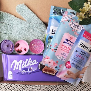 Puede incluir: Una caja de regalo llena de varios productos de ba&ntilde;o y cuerpo, incluyendo bombas de ba&ntilde;o, velas y una barra de chocolate. La caja tambi&eacute;n incluye un par de guantes exfoliantes verdes y un arreglo floral rosa y blanco.
