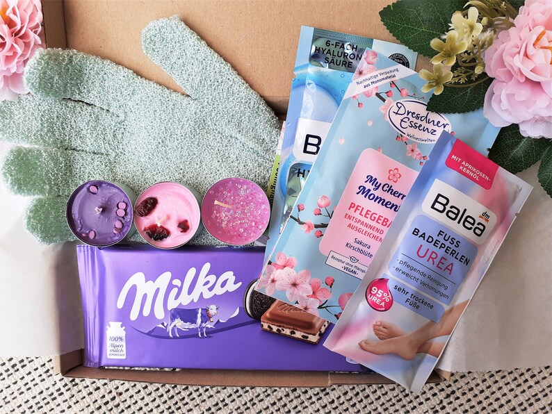 Puede incluir: Una caja de regalo llena de varios productos de ba&ntilde;o y cuerpo, incluyendo bombas de ba&ntilde;o, velas y una barra de chocolate. La caja tambi&eacute;n incluye un par de guantes exfoliantes verdes y un arreglo floral rosa y blanco.