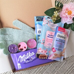 Puede incluir: Una caja de regalo llena de diversos art&iacute;culos de cuidado personal, que incluyen sales de ba&ntilde;o, bombas de ba&ntilde;o, velas, un par de guantes exfoliantes verdes, una barra de chocolate y un ba&ntilde;o de pies. La caja est&aacute; forrada con papel de seda blanco y decorada con una flor rosa.