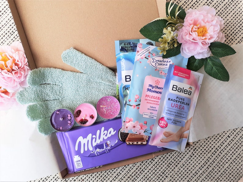 Puede incluir: Una caja de regalo llena de diversos art&iacute;culos de cuidado personal, que incluyen sales de ba&ntilde;o, bombas de ba&ntilde;o, velas, un par de guantes exfoliantes verdes, una barra de chocolate y un ba&ntilde;o de pies. La caja est&aacute; forrada con papel de seda blanco y decorada con una flor rosa.