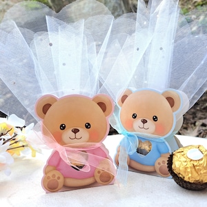 May include: Two teddy bear-shaped gift boxes, one pink and one blue, each with a sheer tulle bow and a heart-shaped cutout. A gold-wrapped chocolate sits nearby. The boxes are light brown with cartoon bear faces.