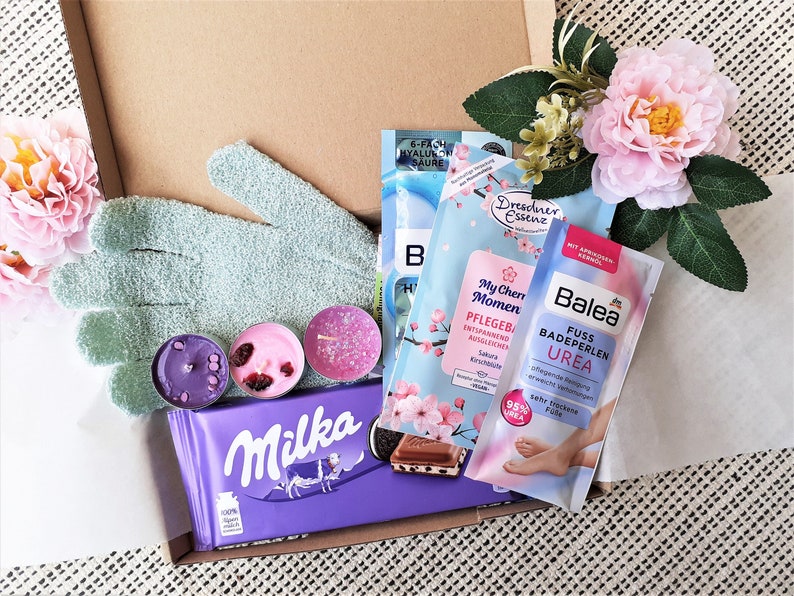 Puede incluir: Una caja de regalo llena de productos de ba&ntilde;o y cuerpo, que incluyen una barra de chocolate morada, un guante exfoliante verde, tres velas peque&ntilde;as y un paquete de producto de ba&ntilde;o rosa y azul. La caja est&aacute; forrada con papel de seda blanco y decorada con flores rosas.