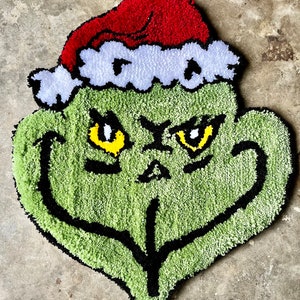 Op de afbeelding: Een groen Grinch-gezichtskleed met een rood-witte kerstmanmuts. De Grinch heeft een ondeugende uitdrukking met gele ogen en een zwarte omtrek.