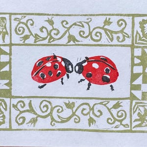 May include: A block print illustration featuring two red ladybugs with black spots positioned in the center. The design is enclosed by a rectangular border in olive green, decorated with floral vines and geometric quilt-like patterns.
