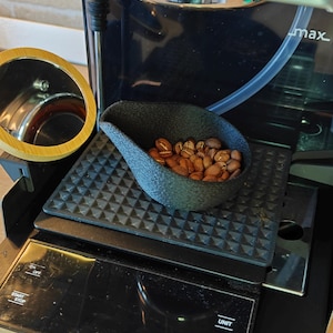 Puede incluir: Una báscula de café negra con una cuchara de granos de café tostados. La báscula tiene botones para las funciones de unidad y tara. Una máquina de espresso Gaggia Classic está en el fondo.