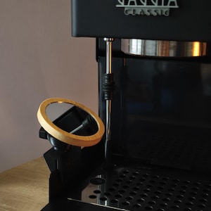 Może przedstawiać: Czarny ekspres do kawy Gaggia Classic ze srebrnym logo. Dołączone jest małe, okrągłe lusterko z drewnianą ramą. Maszyna posiada tackę ociekową ze stali nierdzewnej i chromowaną dyszę do spieniania mleka.