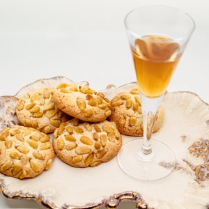 Könnte beinhalten: Ein weißer Keramikteller mit goldenen Akzenten hält ein kleines Glas mit bernsteinfarbener Flüssigkeit und sechs runde Kekse mit Pinienkernen.