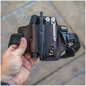 Puede incluir: Una funda de cinturón de cuero marrón con una linterna negra, un bolígrafo y una multiherramienta. La funda tiene un bucle para el cinturón y un clip para sujetarla a un cinturón.