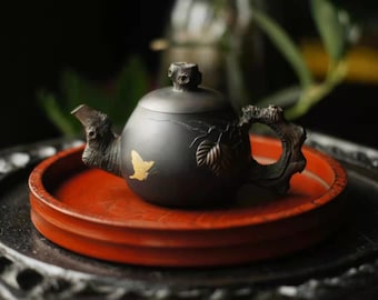 May include: A black teapot with a butterfly design and a wooden handle sits on a red tray. The teapot has a textured surface and a small spout.