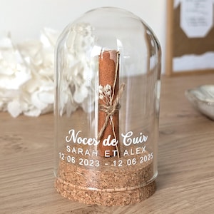 Peut inclure: Un dôme en verre transparent avec une base en liège renferme un parchemin brun roulé, noué avec de la ficelle et des fleurs séchées. Le texte blanc sur le verre indique "Noces de Cuir" avec des noms et des dates. Un souvenir décoratif.