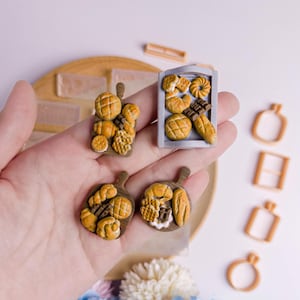 Może przedstawiać: Miniaturowe pieczywo i ciasta na drewnianych deskach do krojenia i blasze do pieczenia. Asortyment obejmuje bochenki, rogaliki i bułki, wszystkie w odcieniach brązu i beżu. Widoczne są również dekoracyjne formy do pieczenia.