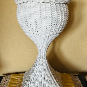 May include: A white wicker pedestal planter featuring a rounded, woven bowl-shaped top section that transitions into a narrow, hourglass-shaped neck, supported by a wide, square-based stand with a consistent woven texture throughout the entire piece.