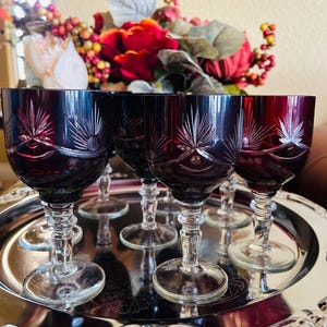 May include: A set of six ruby red wine glasses with etched starburst designs, displayed on a silver serving tray. The glasses have clear stems and bases. A floral arrangement with red roses and berries is in the background.