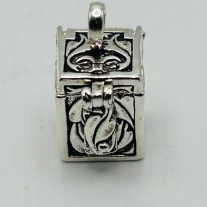 May include: A silver-colored rectangular pendant with a hinged lid and a loop for a chain. The pendant is decorated with floral and abstract designs. The clasp is closed. The pendant is made of metal.