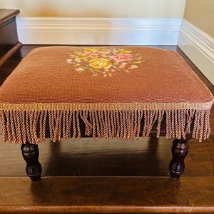 May include: A brown footstool with a floral needlepoint design on top. The footstool has a fringe around the bottom edge and sits on four dark wooden legs. The floral design includes yellow and pink flowers.