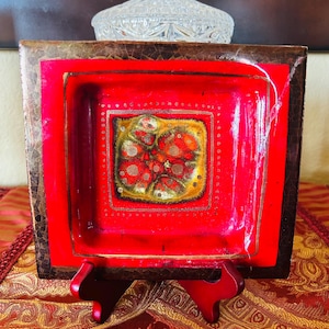 May include: A square, red decorative dish with a brown border. The center of the dish features a floral design in gold, red, and orange. The dish is displayed on a red stand, showcasing its decorative appeal.