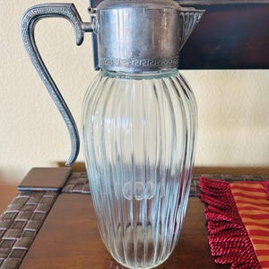May include: A clear glass pitcher with vertical ribbing and a silver-toned metal lid and handle. The metal components feature a decorative Greek key pattern along the rim and handle. The pitcher has a tapered shape and a pointed pouring spout.