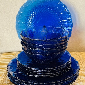 May include: A set of cobalt blue glass dinnerware featuring a stack of plates and a stack of bowls. Each piece has a scalloped edge and a textured leaf pattern embossed on the surface. The glass is translucent and deep blue in color.