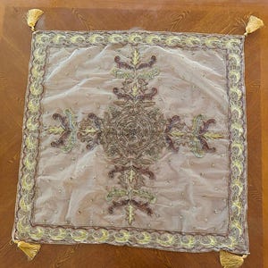 May include: A square, sheer, light brown decorative pillow cover with intricate embroidery in shades of brown, gold, and green. The design features a central circular motif and ornate floral patterns. Gold tassels adorn each corner.