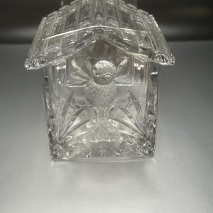 May include: A clear glass birdhouse with a bird figurine on the front. The birdhouse has a detailed design with a roof and windows.