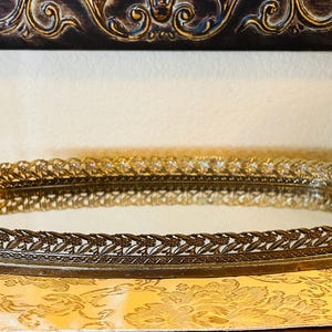May include: An oval, antique-style vanity tray with a mirrored surface. The tray has a decorative, ornate, bronze-colored metal frame with a floral and leaf design. The tray is resting on a yellow floral patterned surface.