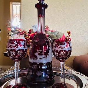 May include: A ruby red glass decanter with a matching stopper and two wine glasses. The decanter and glasses feature etched deer and forest scenes. The set is displayed on a silver tray.