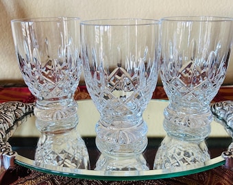 May include: Three clear crystal glasses with diamond-cut patterns are displayed on a mirrored tray with ornate silver edges. The glasses have a short, rounded base and a wide, straight rim.