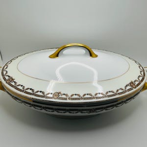 May include: White oval serving dish with a lid, gold handles, and decorative brown and gold trim. The dish has a gold handle on the lid and gold handles on each side. The dish is likely made of porcelain.
