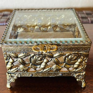 May include: A gold-toned metal trinket box with a glass top and intricate rose design. The box has four feet and a hinged lid with a decorative knob.