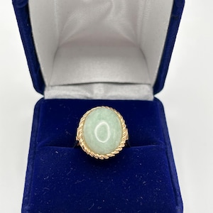 May include: A gold ring with a large, oval, pale green stone. The ring is set in a gold band with a twisted rope design around the stone. The ring is displayed in a blue velvet ring box.