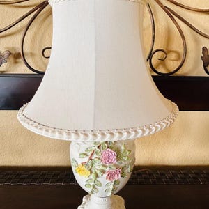 May include: A table lamp with a cream-colored fabric shade featuring a braided trim and a pink rose finial. The base is white with a sculpted floral design of pink and yellow roses with green leaves, set upon a decorative pedestal base.