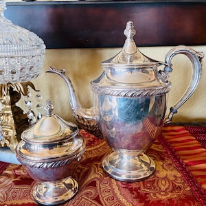 May include: A silver tea set with a teapot and a sugar bowl. The teapot has a curved spout and a handle, and the sugar bowl has a lid. Both pieces are on a red and gold patterned tablecloth.