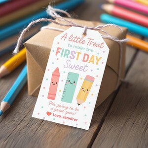 May include: A gift box wrapped in brown paper with a tag that reads "A little Treat to make the FIRST DAY Sweet." The tag features illustrations of a crayon, ruler, and pencil. The text "It's going to be a great year! Love, Jennifer" is also on the tag.