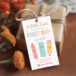 May include: A gift tag with the text "A little Treat to make the FIRST DAY Sweet." The tag features illustrations of a crayon, ruler, and pencil. The tag is tied to a wrapped gift with twine.