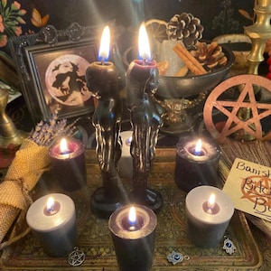 May include: A collection of lit candles, including two black figure candles, surrounded by smaller candles in various colors. A tray holds the candles, along with a pentagram, a framed picture, and other mystical items. The background features a celestial-themed tapestry.