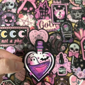May include: A collection of gothic-themed stickers in shades of pink, purple, and black. One sticker features a heart-shaped bottle with a ghost and bat inside. Other stickers include skulls, a black cat, and the text "Got Queen".