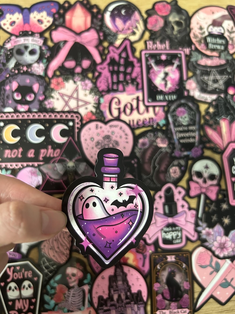 May include: A collection of gothic-themed stickers in shades of pink, purple, and black. One sticker features a heart-shaped bottle with a ghost and bat inside. Other stickers include skulls, a black cat, and the text "Got Queen".