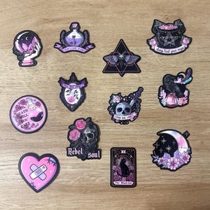 May include: A collection of gothic-themed stickers featuring various designs. Includes a crystal ball, potion bottles, a moth, a cauldron with a pentagram, a raven, a skull with roses, a heart with a bandage, and a tarot card with a black cat. The stickers have a dark color palette.