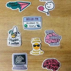 May include: A collection of colorful stickers with humorous phrases and graphics. Designs include a paper airplane, a brain, an alien, a shopping bag, a computer, and an emoji wearing sunglasses. Text includes "Kindly F*ck Off", "Hello I'm Allergic to People", and "Let Me Overthink This."